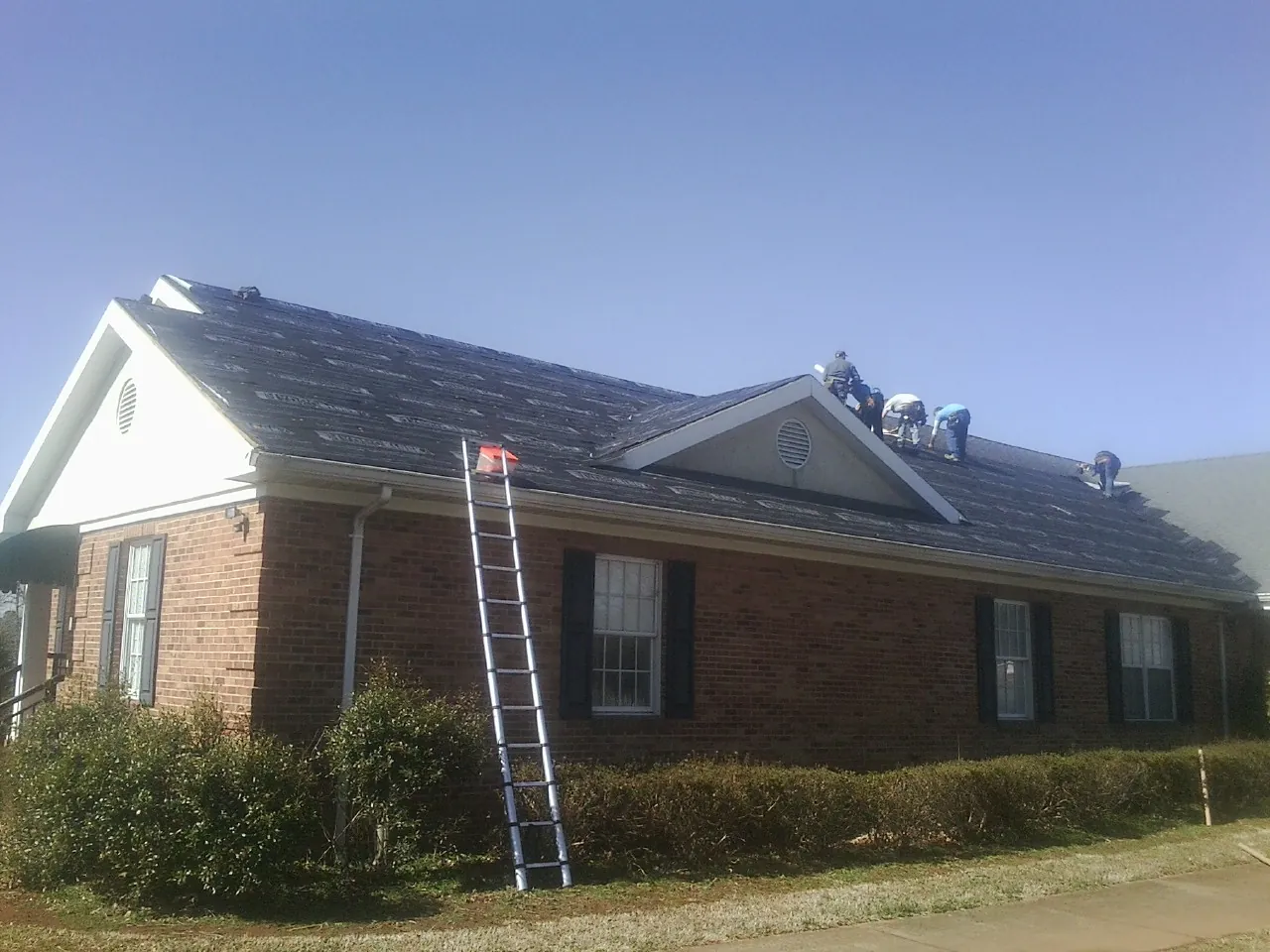 Crew tearing shingles off a brick ranch roof with a ladder set — Metal Roof Repair work by our crew in Sunfield